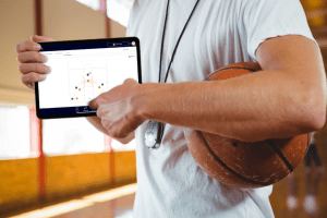 Basketball coach holding a tablet displaying a basketball plays diagram on screen while carrying a basketball and whistle in a gym.